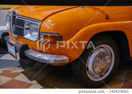 Detailed close-up view of front corner of vintage orange car. Highlights include shiny grille, headlight, chrome bumper, and a portion of the tire and wheel against a checkered floor background. Detailed close-up view of front corner of vintage orange car. Highlights include shiny grille, headlight, chrome bumper, and a portion of the tire and wheel against a checkered floor background. 140459081