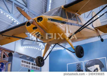 Vintage yellow propeller airplane suspended inside an aviation museum with bright interior lighting and industrial architecture. Historic aircraft with classic wings and landing gear displayed as part Vintage yellow propeller airplane suspended inside an aviation museum with bright interior lighting and industrial architecture. Historic aircraft with classic wings and landing gear displayed as part 140459085
