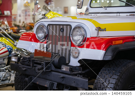 Close-up view of vintage white off-road vehicle featuring bold red and yellow racing stripes, chrome grille, and round headlights. Partially visible is a custom motorcycle with a star-spangled fender 140459105