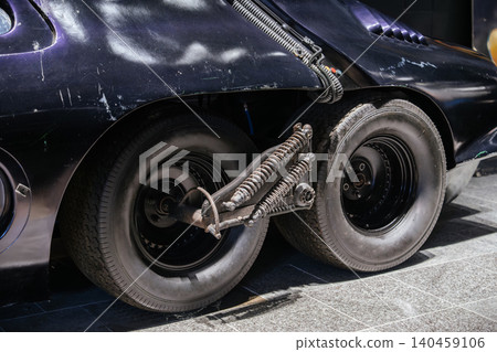 Detailed close-up captures rear suspension and dual tires of custom vehicle. Complex mechanical components and worn dark body highlight specialized build under bright daylight. 140459106