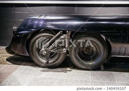 Close-up view of custom vehicle's rear wheel assembly showcasing twin tires and advanced suspension system. Dark glossy bodywork features stylized vents against a tiled floor and brick wall. 140459107