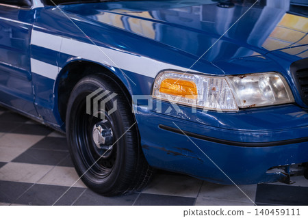 Close-up, low-angle view captures front corner of blue sedan with white stripes. Vehicle shows signs of age with scratches and rust, set against a checkered garage floor. 140459111