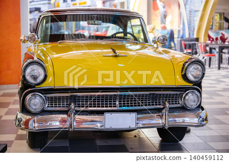 Close-up shot of a vintage yellow car with a dark roof, showcasing chrome details. The car is parked indoors on a checkered floor, with a blurred diner background featuring red-seated tables and 140459112