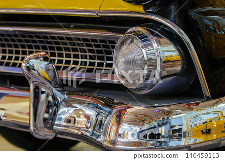 Detailed close-up captures front end of vintage car, showcasing polished chrome bumper, checkered grille, and glass headlight with chrome accents. Reflective surfaces mirror classic automobiles and Detailed close-up captures front end of vintage car, showcasing polished chrome bumper, checkered grille, and glass headlight with chrome accents. Reflective surfaces mirror classic automobiles and 140459113