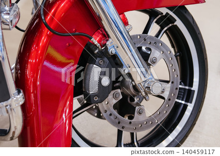 Detailed close-up captures front wheel assembly of motorcycle. Vibrant metallic red fender curves over polished chrome front fork, showcasing mechanical components against a blurred background. Detailed close-up captures front wheel assembly of motorcycle. Vibrant metallic red fender curves over polished chrome front fork, showcasing mechanical components against a blurred background. 140459117