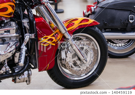 Close-up shot features custom motorcycle's front wheel and fender, showcasing vibrant red color with yellow and orange flame graphics. Polished chrome details enhance the striking design, with another 140459119
