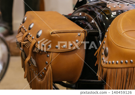 Close-up of a brown leather motorcycle saddlebag featuring decorative metal studs and fringes. A portion of a matching motorcycle seat is visible, showcasing craftsmanship and vintage style. Close-up of a brown leather motorcycle saddlebag featuring decorative metal studs and fringes. A portion of a matching motorcycle seat is visible, showcasing craftsmanship and vintage style. 140459126