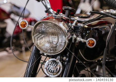 Detailed close-up photograph showcases iconic front of vintage motorcycle. Focus on large circular headlight with ribbed glass lens and chrome trim, flanked by amber turn signals. Detailed close-up photograph showcases iconic front of vintage motorcycle. Focus on large circular headlight with ribbed glass lens and chrome trim, flanked by amber turn signals. 140459130