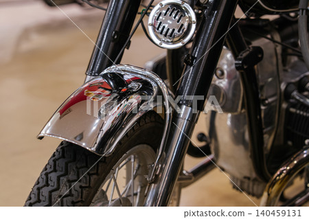 Detailed close-up view of vintage motorcycle front end, showcasing shiny chrome fender, black tire with defined tread, and visible wheel spokes. A round metallic component, possibly a horn, is mounted Detailed close-up view of vintage motorcycle front end, showcasing shiny chrome fender, black tire with defined tread, and visible wheel spokes. A round metallic component, possibly a horn, is mounted 140459131