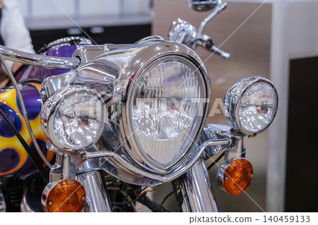 Detailed close-up view of front of custom motorcycle, showcasing chrome components and lighting system. Central headlight is prominent, flanked by smaller lights and striking flame graphic. 140459133