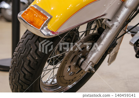 Detailed view captures motorcycle's front wheel, spoked rim, and bright yellow fender with chrome trim. Amber turn signal and worn tire add character, showcasing mechanical design in a blurred indoor Detailed view captures motorcycle's front wheel, spoked rim, and bright yellow fender with chrome trim. Amber turn signal and worn tire add character, showcasing mechanical design in a blurred indoor 140459141