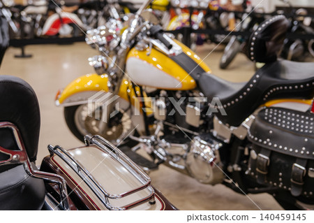 Close-up shot features a striking yellow and white motorcycle with polished chrome accents and a black studded seat. Background displays blurred motorcycles in a bright dealership setting. 140459145