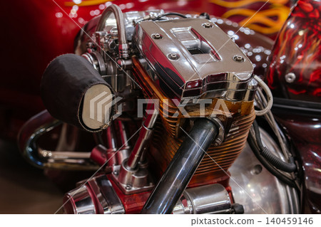 Detailed close-up of a custom V-twin motorcycle engine featuring a shiny chrome valve cover and copper-colored cooling fins. Background shows blurred reflections of a vehicle body with vibrant flame Detailed close-up of a custom V-twin motorcycle engine featuring a shiny chrome valve cover and copper-colored cooling fins. Background shows blurred reflections of a vehicle body with vibrant flame 140459146