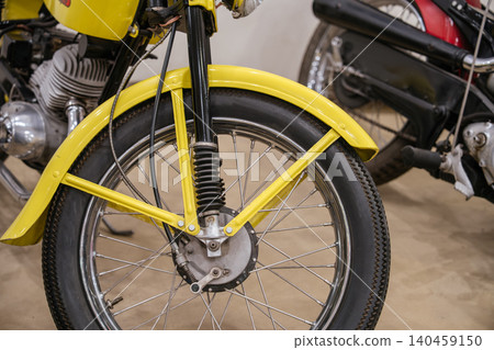 Detailed close-up captures front wheel and suspension of vintage yellow motorcycle. Vibrant yellow fender contrasts with black fork tubes and chrome spokes, showcasing classic design elements. Detailed close-up captures front wheel and suspension of vintage yellow motorcycle. Vibrant yellow fender contrasts with black fork tubes and chrome spokes, showcasing classic design elements. 140459150