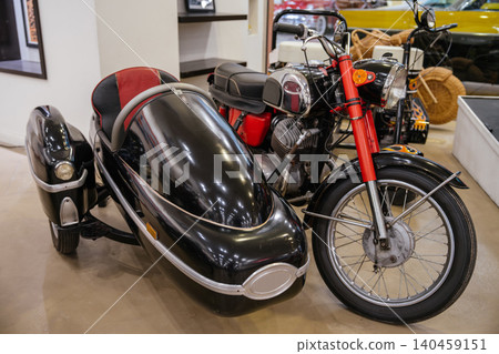 Classic motorcycle with a black sidecar displayed indoors. The motorcycle features a red frame and chrome details, while the sidecar boasts a glossy finish and red-black upholstery, creating a Classic motorcycle with a black sidecar displayed indoors. The motorcycle features a red frame and chrome details, while the sidecar boasts a glossy finish and red-black upholstery, creating a 140459151