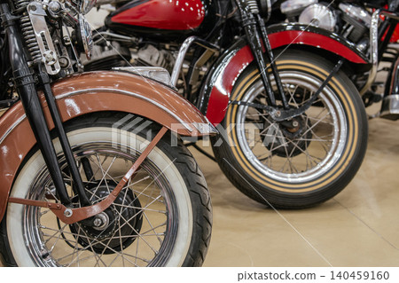 Detailed view captures front wheels and fenders of two vintage motorcycles parked indoors. Foreground motorcycle features a copper fender and whitewall tire, while background motorcycle showcases a 140459160