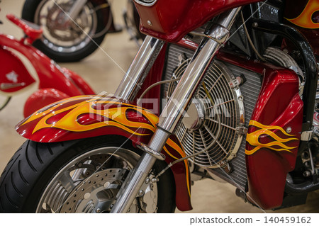 Detailed close-up captures front section of custom motorcycle. Striking metallic red paint features intricate airbrushed flame graphics in vibrant yellow and orange, showcasing polished chrome 140459162