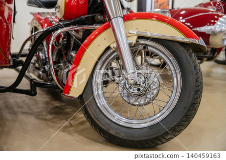 Detailed low-angle view captures front wheel and two-tone cream and red fender of classic motorcycle. Chrome accents and deep-tread tire enhance vintage appeal against polished floor. Detailed low-angle view captures front wheel and two-tone cream and red fender of classic motorcycle. Chrome accents and deep-tread tire enhance vintage appeal against polished floor. 140459163