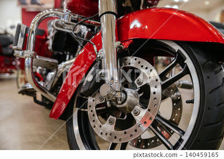 Detailed view highlights front wheel, tire, brake disc, and chrome components of red motorcycle. Low-angle perspective emphasizes robust engineering and polished surfaces in showroom setting. Detailed view highlights front wheel, tire, brake disc, and chrome components of red motorcycle. Low-angle perspective emphasizes robust engineering and polished surfaces in showroom setting. 140459165
