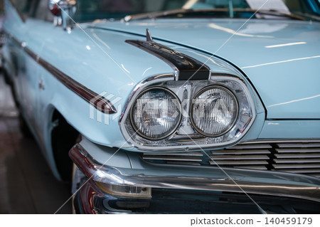 Detailed frontal view of classic pale blue automobile, showcasing dual round headlights, chrome grille, and shiny chrome bumper. Decorative chrome trim and hood ornament enhance vintage appeal. Detailed frontal view of classic pale blue automobile, showcasing dual round headlights, chrome grille, and shiny chrome bumper. Decorative chrome trim and hood ornament enhance vintage appeal. 140459179