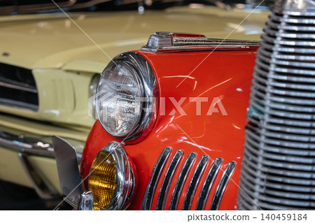 Detailed close-up photograph showcases front-end design of two vintage cars. A vibrant orange car with chrome accents stands out, while a pale yellow car is softly blurred in the background Detailed close-up photograph showcases front-end design of two vintage cars. A vibrant orange car with chrome accents stands out, while a pale yellow car is softly blurred in the background 140459184