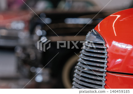 Detailed view highlights polished chrome grille and vibrant orange fender of vintage automobile. Softly blurred background reveals another classic car, enhancing automotive nostalgia. 140459185