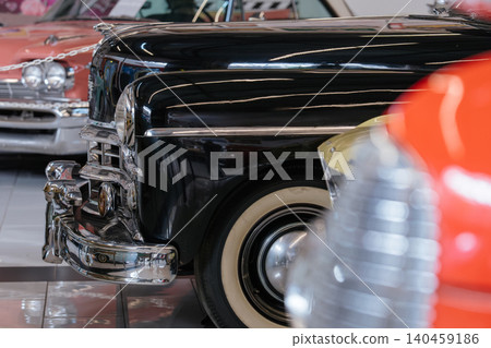 Close-up shot highlights vintage black car's chrome grille, bumper, and whitewall tire. Other classic cars, including pink and red models, are visible in the polished showroom background. Close-up shot highlights vintage black car's chrome grille, bumper, and whitewall tire. Other classic cars, including pink and red models, are visible in the polished showroom background. 140459186