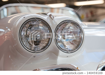 Close-up view highlights dual round headlights of a classic white automobile. Ribbed glass lenses and chrome bezels reflect ambient light, showcasing vintage automotive design in a garage setting. Close-up view highlights dual round headlights of a classic white automobile. Ribbed glass lenses and chrome bezels reflect ambient light, showcasing vintage automotive design in a garage setting. 140459189
