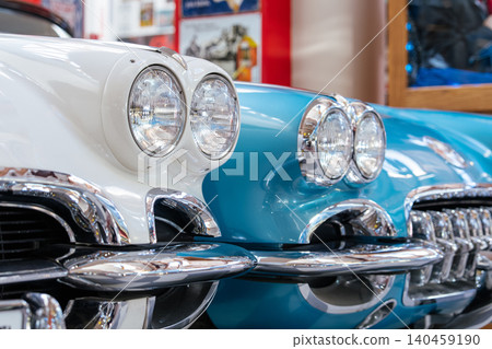 Detailed close-up shot features two vintage American sports cars, one white and the other turquoise, showcasing iconic headlights and chrome grilles. Bright lighting highlights polished surfaces 140459190