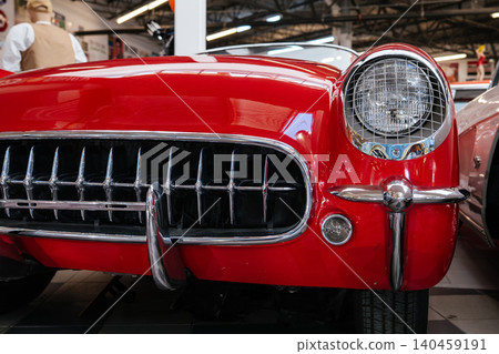 Detailed low-angle view highlights front grille and headlight of vibrant red classic convertible sports car. Chrome details gleam under showroom lighting, showcasing vintage automotive design. 140459191