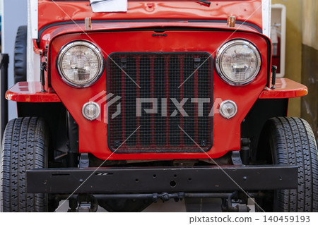 Close-up view of classic red off-road vehicle showcasing round chrome-rimmed headlights and vertical slatted grille. Sturdy black bumper and large treaded tires enhance rugged appearance, suggesting Close-up view of classic red off-road vehicle showcasing round chrome-rimmed headlights and vertical slatted grille. Sturdy black bumper and large treaded tires enhance rugged appearance, suggesting 140459193