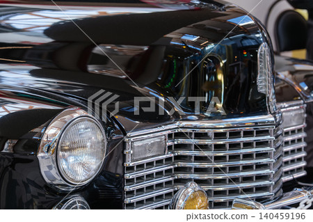 Detailed view of front of classic black car, showcasing polished chrome grille and round headlight. Gleaming exterior reflects indoor exhibition space, highlighting vintage design and craftsmanship. 140459196