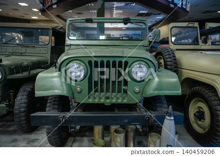 Collection of olive-drab military jeeps parked closely together in a museum. Central jeep showcases classic features like a seven-slot grille and round headlights, with artillery shells in the Collection of olive-drab military jeeps parked closely together in a museum. Central jeep showcases classic features like a seven-slot grille and round headlights, with artillery shells in the 140459206