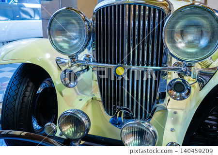 Detailed view of front section of classic pale yellow vintage automobile. Intricate chrome grille, dual round headlights, and auxiliary light shine brightly in exhibition setting. Detailed view of front section of classic pale yellow vintage automobile. Intricate chrome grille, dual round headlights, and auxiliary light shine brightly in exhibition setting. 140459209