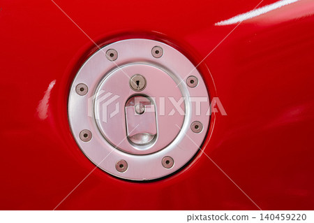 Detailed view of a metallic fuel cap assembly on a shiny red vehicle. The circular cap features screws, a keyhole, and a flap, highlighting automotive design and vibrant color. Detailed view of a metallic fuel cap assembly on a shiny red vehicle. The circular cap features screws, a keyhole, and a flap, highlighting automotive design and vibrant color. 140459220
