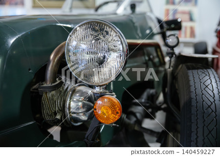 Detailed view captures front section of vintage green car. Focus on large circular headlight with chrome trim, amber turn signal, and robust tire showcasing distinct tread pattern. Detailed view captures front section of vintage green car. Focus on large circular headlight with chrome trim, amber turn signal, and robust tire showcasing distinct tread pattern. 140459227