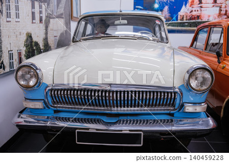 Frontal view of a vintage two-tone white and light blue sedan displayed indoors. The car features a chrome grille and round headlights, with part of the interior visible alongside a vibrant city mural Frontal view of a vintage two-tone white and light blue sedan displayed indoors. The car features a chrome grille and round headlights, with part of the interior visible alongside a vibrant city mural 140459228