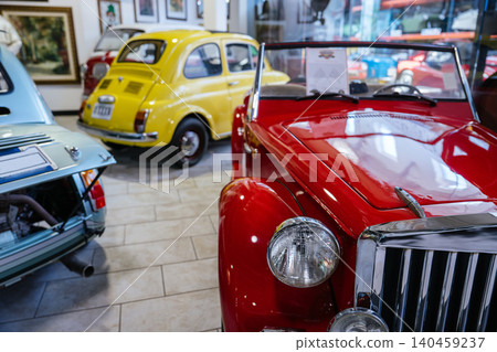 Detailed view of front of shiny red vintage convertible sports car with chrome grille and round headlight. Displayed indoors, surrounded by other vintage cars in a bright showroom environment. Detailed view of front of shiny red vintage convertible sports car with chrome grille and round headlight. Displayed indoors, surrounded by other vintage cars in a bright showroom environment. 140459237