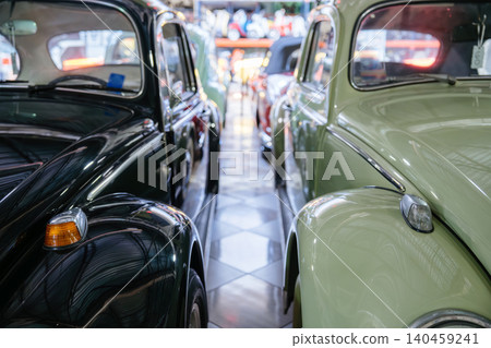 Close-up view highlights front fenders and headlights of two vintage cars parked side by side. Dark glossy car contrasts with pale green vehicle, showcasing classic design in a nostalgic showroom 140459241