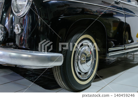 Detailed low-angle view captures front corner of classic black car. Shiny chrome bumper and reflective surfaces highlight automotive elegance in an indoor setting. Detailed low-angle view captures front corner of classic black car. Shiny chrome bumper and reflective surfaces highlight automotive elegance in an indoor setting. 140459246