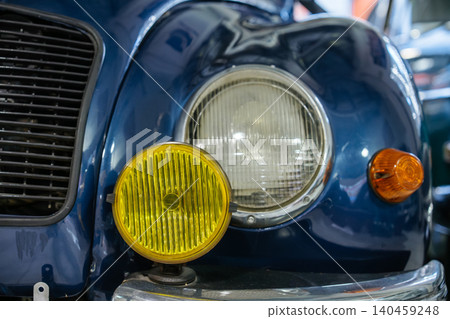 Close-up shot captures front of vintage blue car, showcasing large round headlight with chrome bezel and yellow auxiliary light. Glossy paint reflects surroundings, hinting at indoor setting. Close-up shot captures front of vintage blue car, showcasing large round headlight with chrome bezel and yellow auxiliary light. Glossy paint reflects surroundings, hinting at indoor setting. 140459248