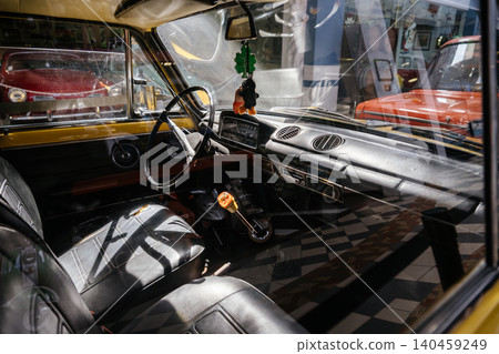 Interior view captures vintage car's dashboard, steering wheel, and gear shift. Sunlight highlights details like the checkered floor and reflections of other classic cars outside. 140459249