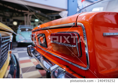 Detailed view captures rear of shiny orange vintage car, emphasizing red taillight and chrome bumper. Soft-focus background features classic cars, set in a bright garage with checkered floor. Detailed view captures rear of shiny orange vintage car, emphasizing red taillight and chrome bumper. Soft-focus background features classic cars, set in a bright garage with checkered floor. 140459252