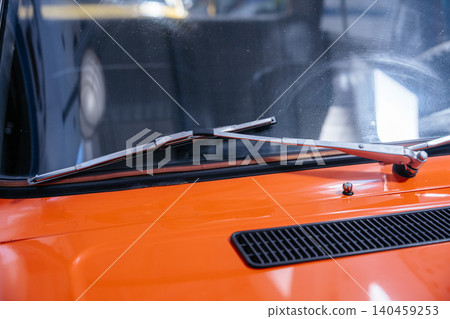 Close-up view captures metal and rubber windshield wipers on a dusty windshield of an orange vehicle. Vibrant orange hood features a black ventilation grate, with a blurred wheel reflected. 140459253