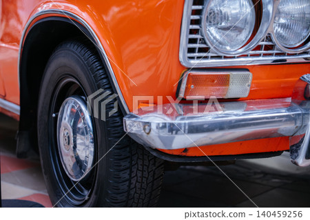 Detailed close-up captures front corner of vintage orange car. Shiny paint, chrome hubcap, tire, headlight, and bumper highlight classic design and reflective surfaces. Detailed close-up captures front corner of vintage orange car. Shiny paint, chrome hubcap, tire, headlight, and bumper highlight classic design and reflective surfaces. 140459256