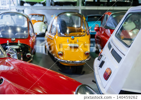 Collection of vintage microcars displayed in a well-lit indoor space. Bright red hood and fender dominate foreground, while a vibrant yellow microcar and a white rear are also visible. 140459262
