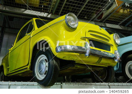 Bright yellow vintage car showcased from low angle, highlighting chrome accents and unique headlights. Positioned on metal platform in what appears to be a collector's exhibition area with another 140459265