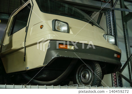 Close-up view of a vintage three-wheeled microcar in a storage facility. The light beige vehicle features a dark grey bumper and integrated headlights, with metal shelving units in the background. Close-up view of a vintage three-wheeled microcar in a storage facility. The light beige vehicle features a dark grey bumper and integrated headlights, with metal shelving units in the background. 140459272