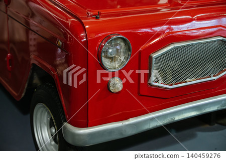 Detailed close-up view of front portion of classic red vehicle. Glossy paint reflects light, showcasing contours. Features round headlight with chrome bezel and metallic mesh grille. 140459276