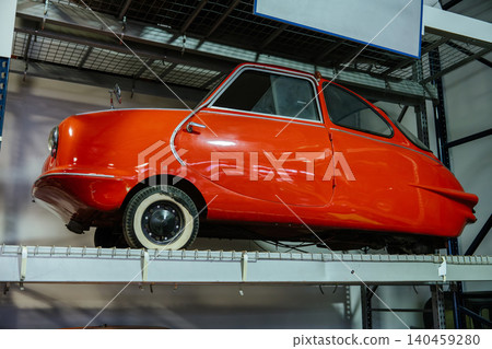 Vibrant orange vintage three-wheeled microcar showcased on a metal shelf in a museum-like setting. The car features a rounded design, chrome accents, and whitewall tires, illuminated by bright 140459280